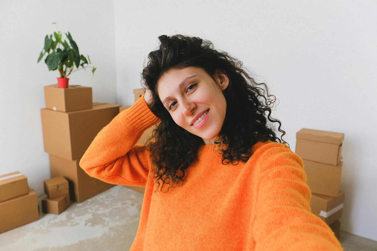 Cheerful young female smiling and touching curly hair while taking selfie against pile of cardboard boxes after relocation in new apartment