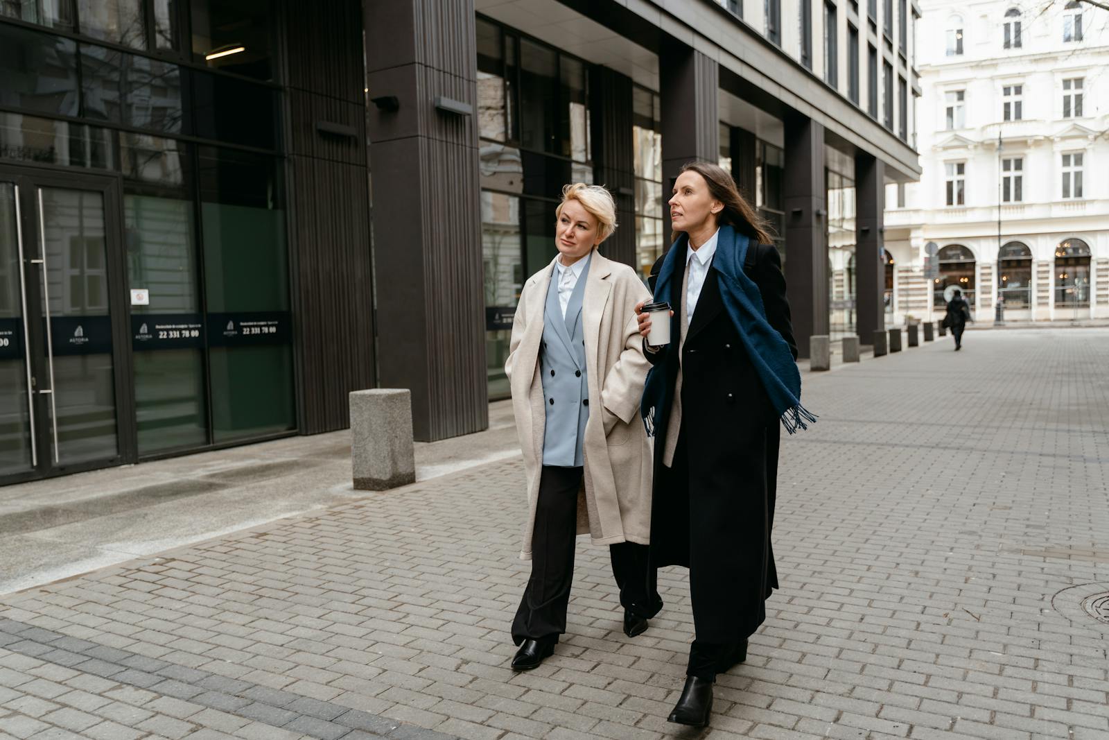 Two businesswomen in formal attire walking and talking outdoors with coffee cups.