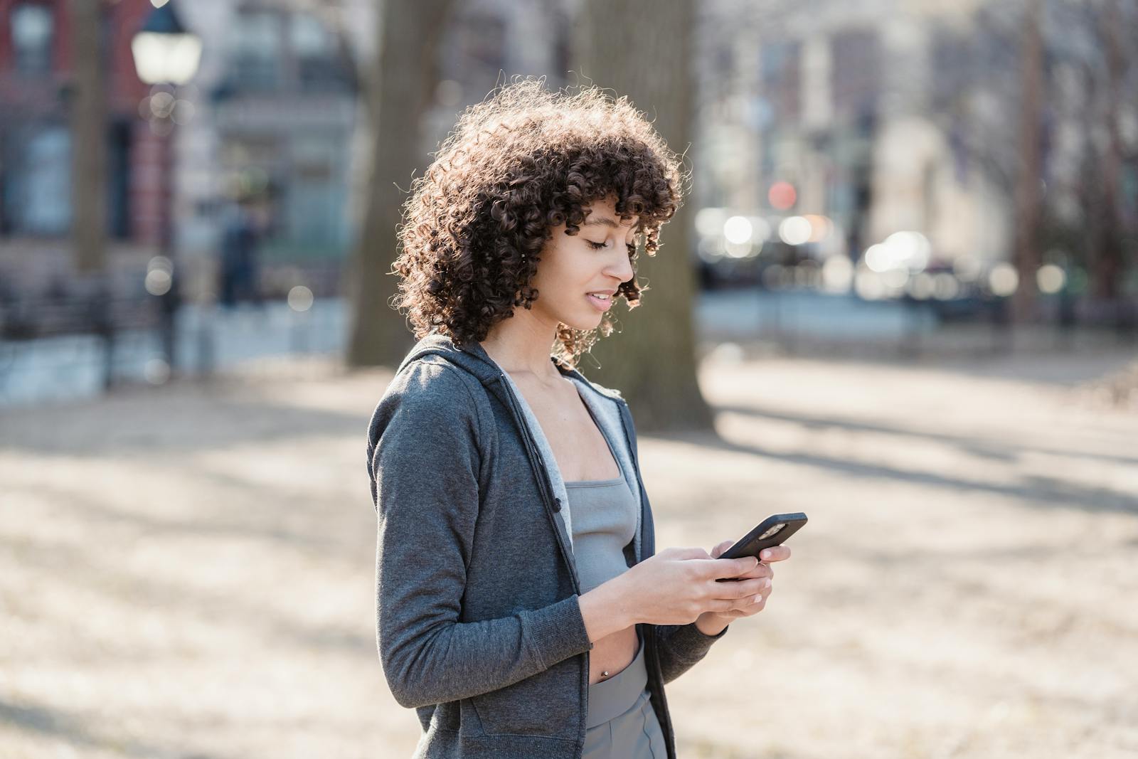 African American female in gray outfit typing text message while walking in park against blurred background