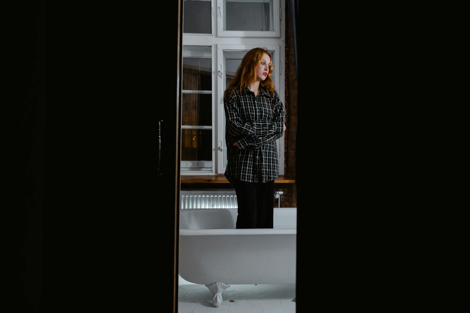 Woman in a flannel shirt standing by a bathtub, reflecting alone.