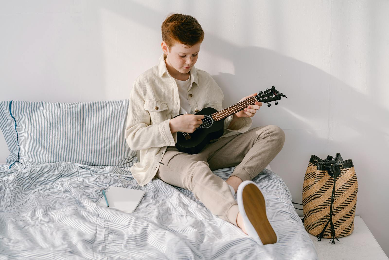 Preteen boy plays ukulele on a cozy bed, creating a serene and artistic atmosphere.