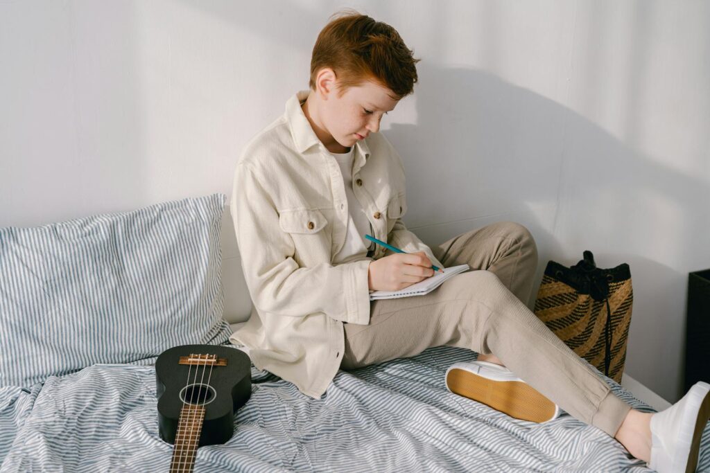 Preteen boy sitting on bed jotting down notes with a ukulele beside him, cozy indoor setting.