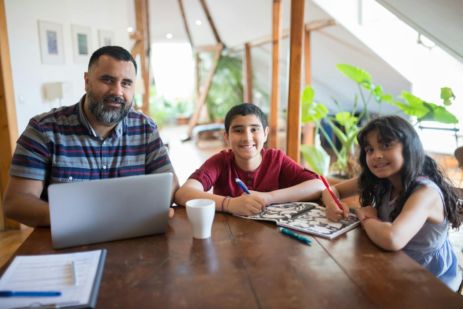 A father happily spends time with his son and daughter at home, engaged in drawing and using a laptop.