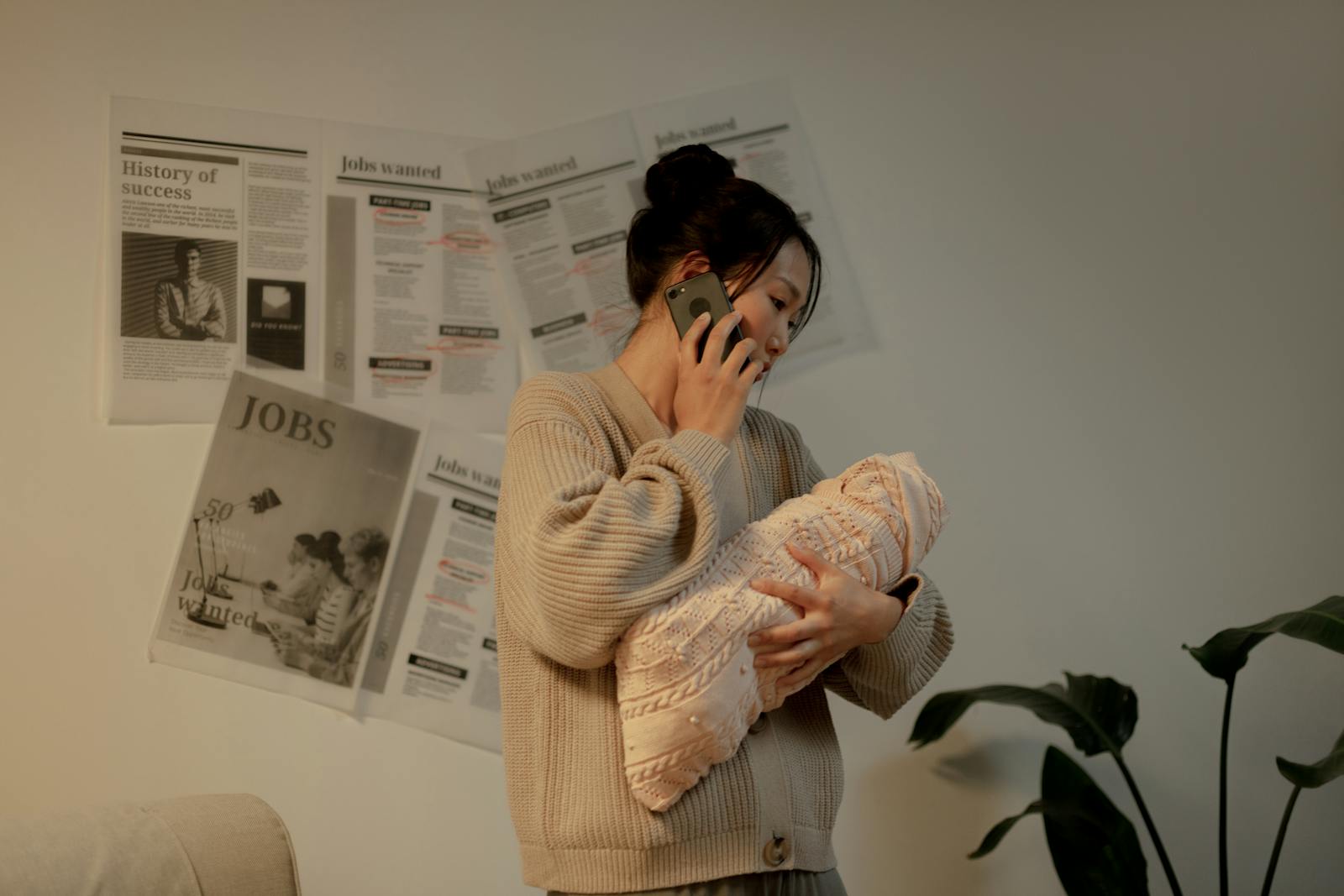 A mother multitasking with her newborn and phone indoors, surrounded by job ads on the wall.