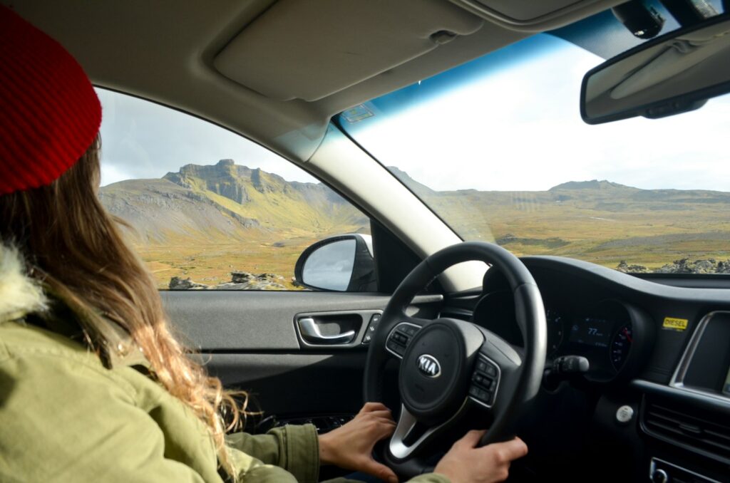 a woman driving a car on a road with mountains in the background