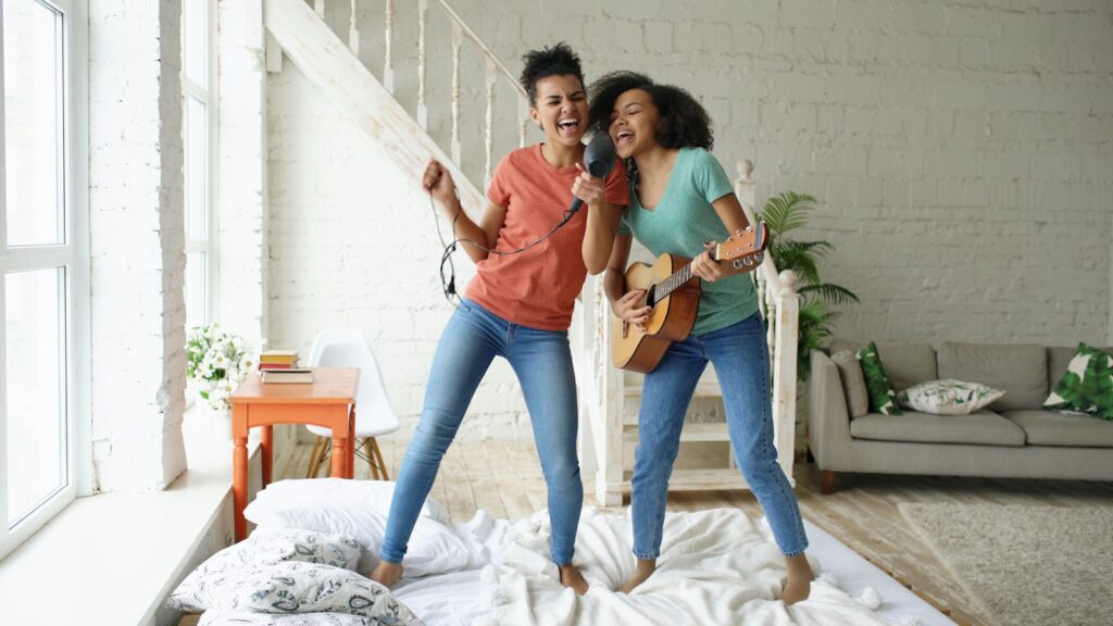 Two women singing and playing guitar on a bed.