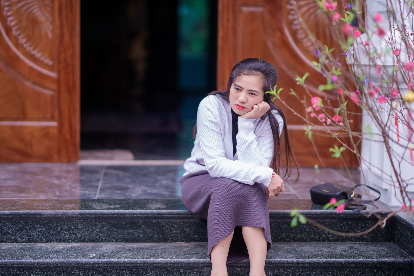 Young woman in a skirt sits thoughtfully on steps beside flowering branches and a wooden doorway.