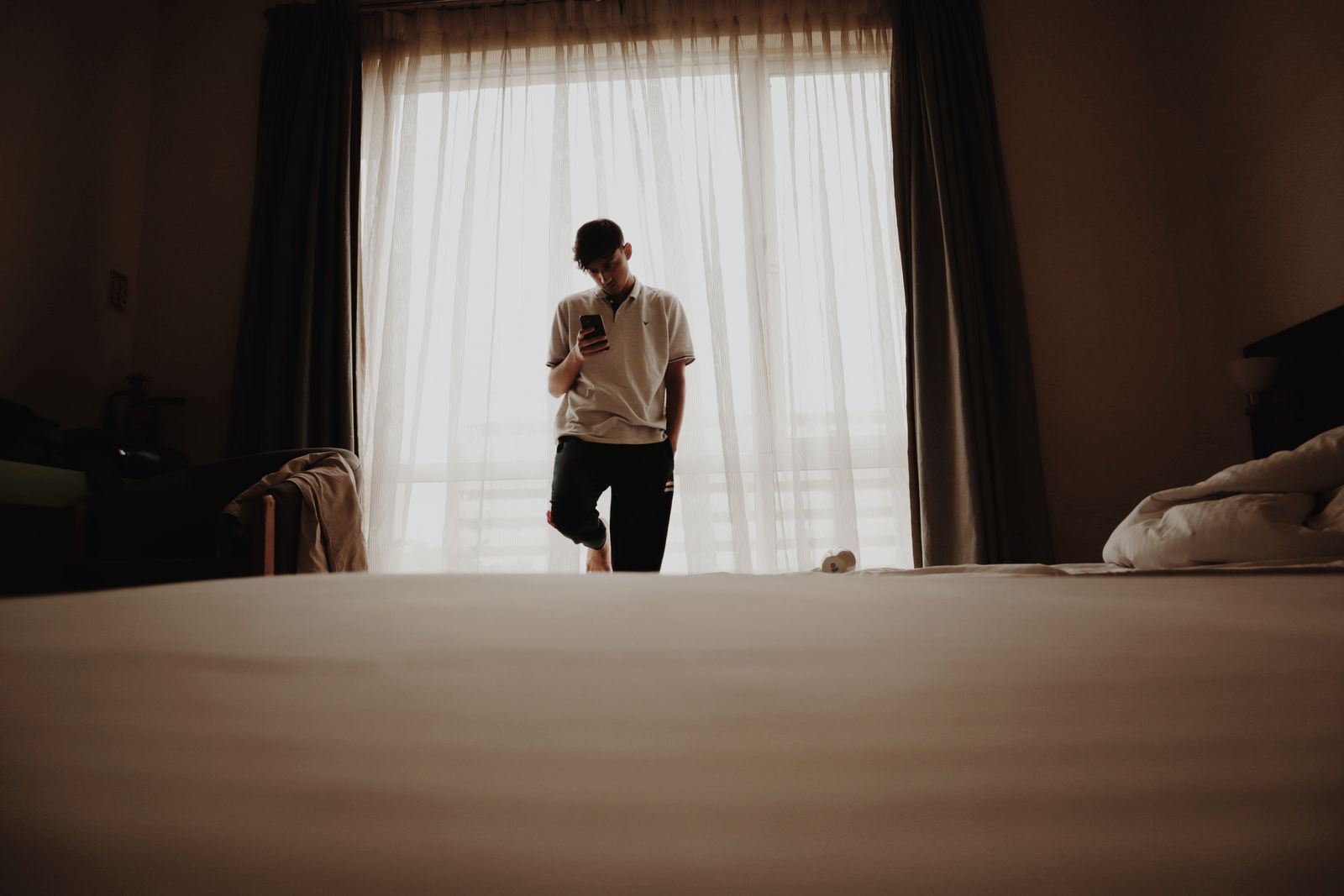 Young man in a polo shirt stands by a bedroom window browsing on his smartphone.