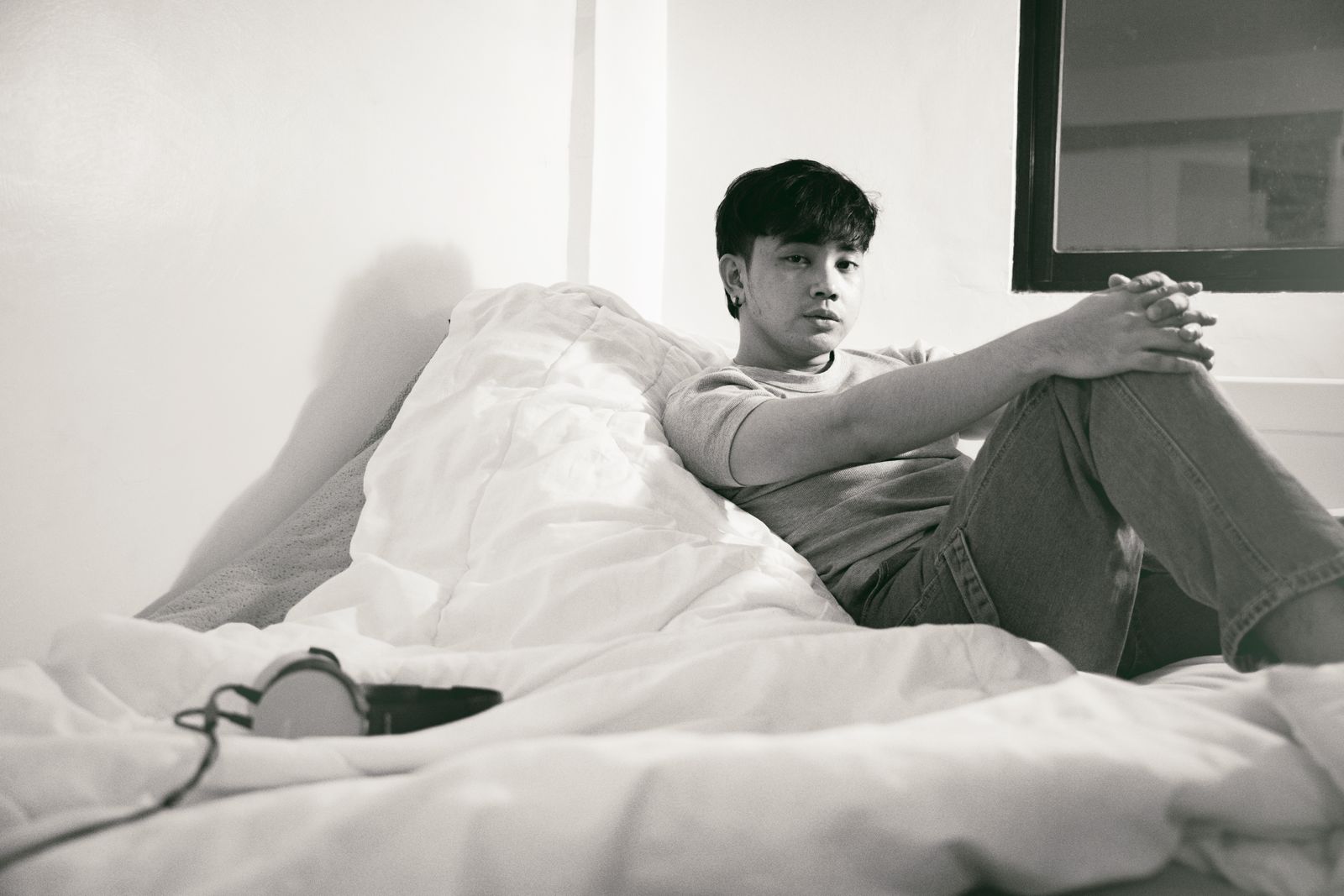 A young man in casual wear sits on a bed in a relaxed pose. Black and white interior portrait.