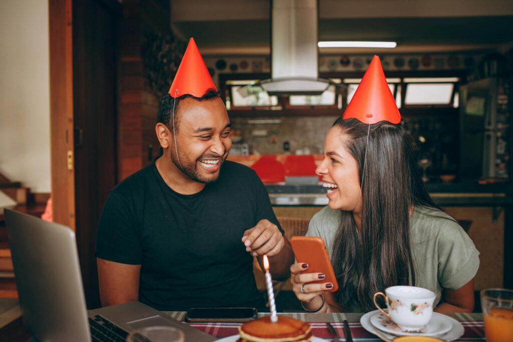Two friends celebrating a birthday indoors with party hats, a cake, and laughter.