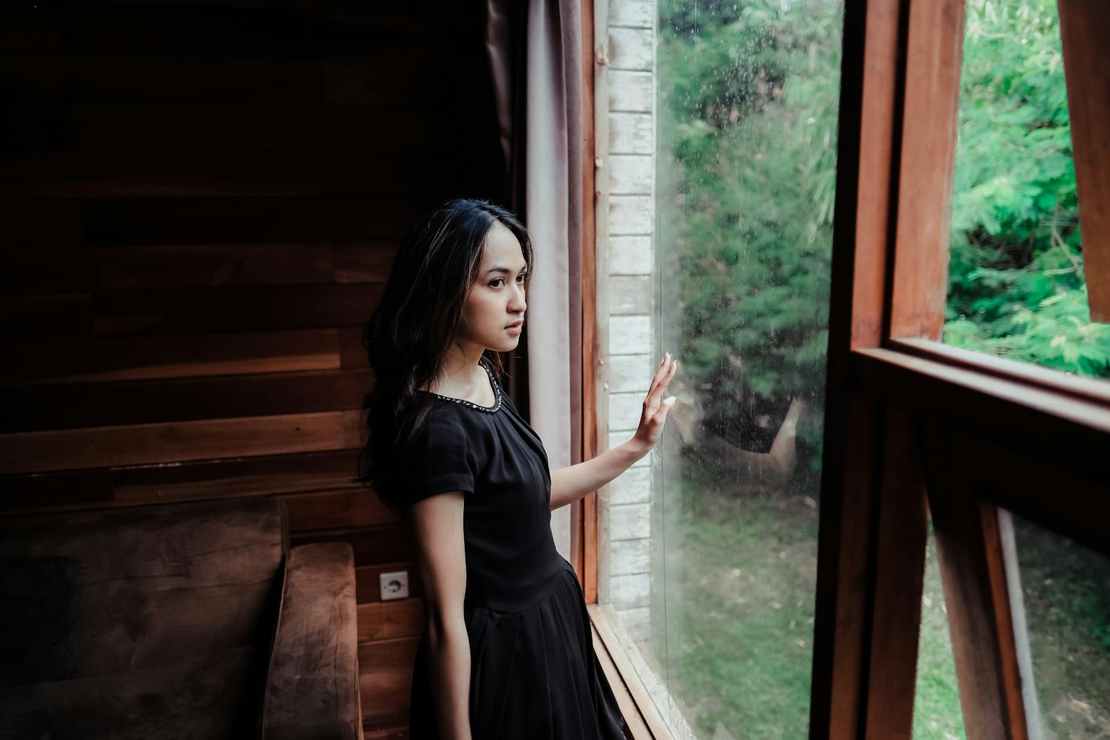 A thoughtful woman in a black dress gazes out a wooden window, exuding elegance and mystery.