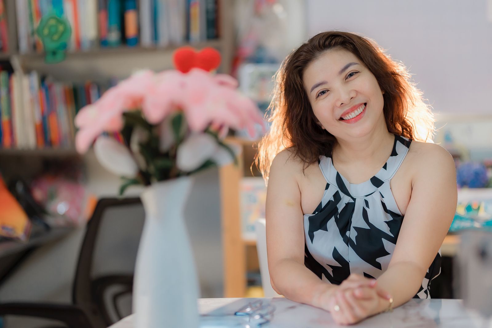 A cheerful woman smiling while seated at a desk in a cozy home environment, surrounded by books.