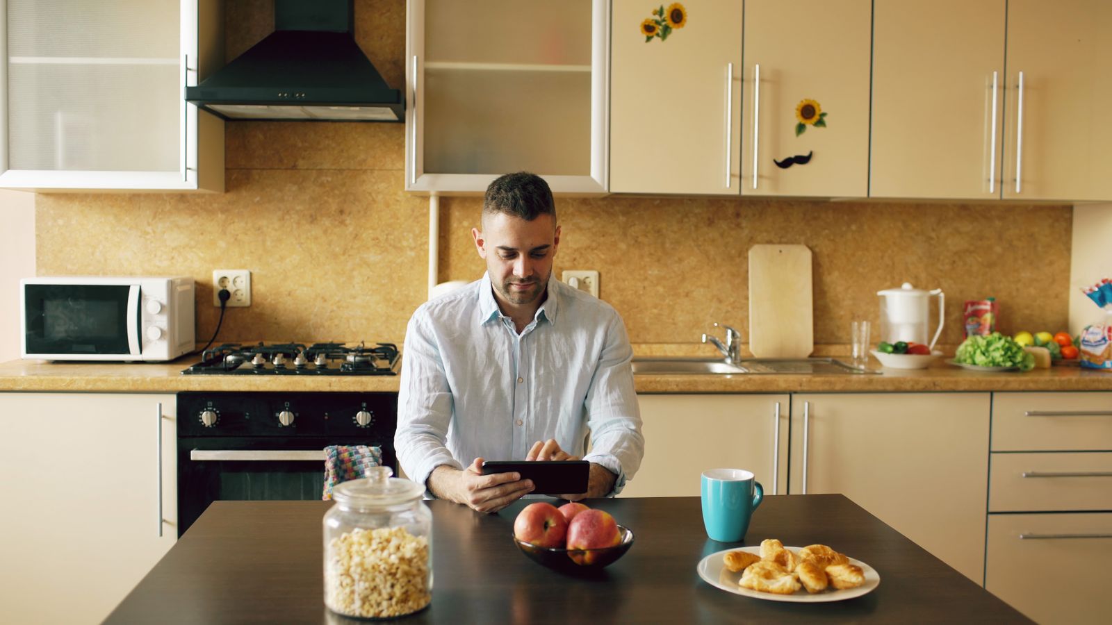 Man sitting at kitchen table using a tablet, surrounded by breakfast items and modern kitchen appliances.