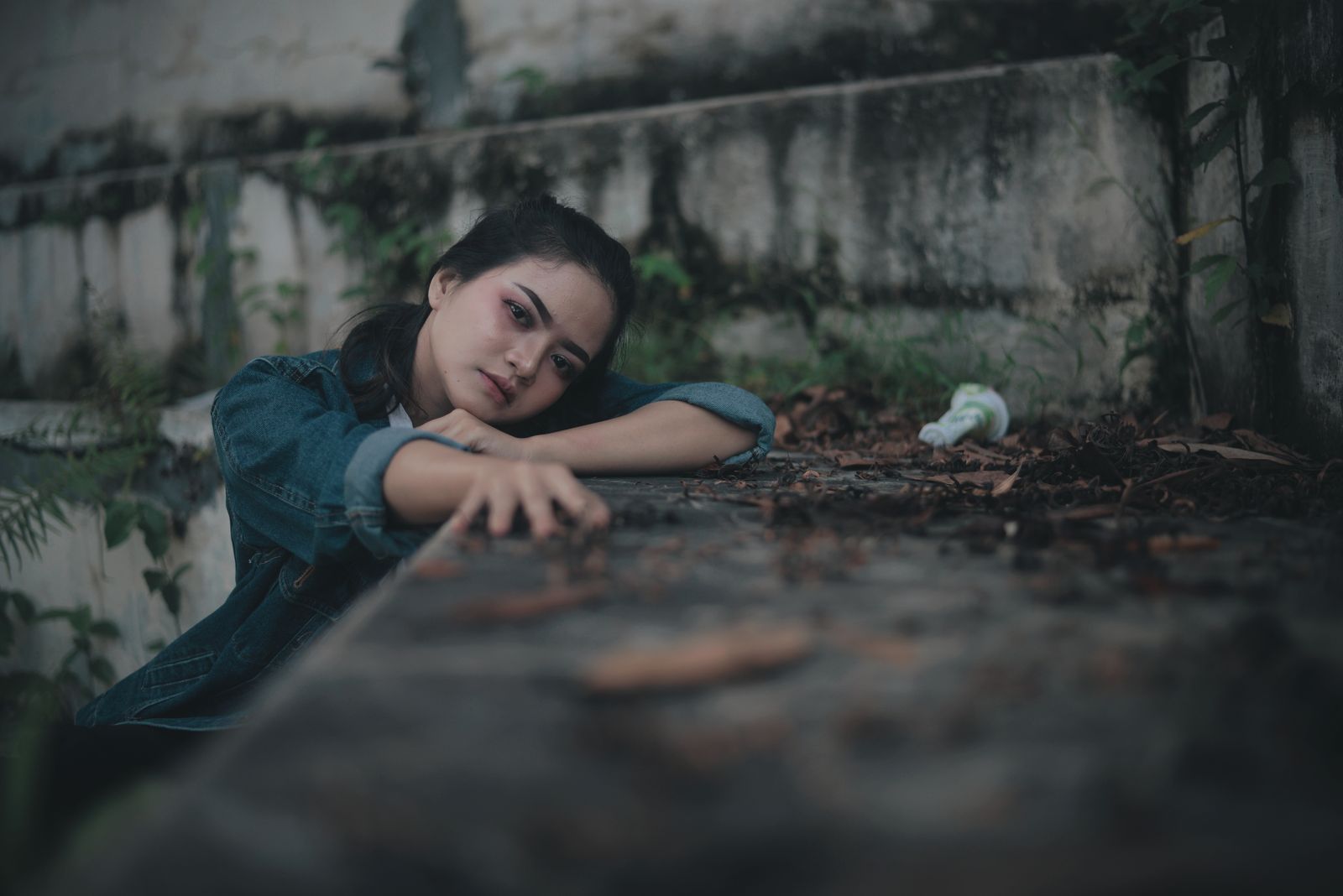 Sad pensive ethnic female teenager in jeans jacket looking at camera while sitting alone near old stone wall in park