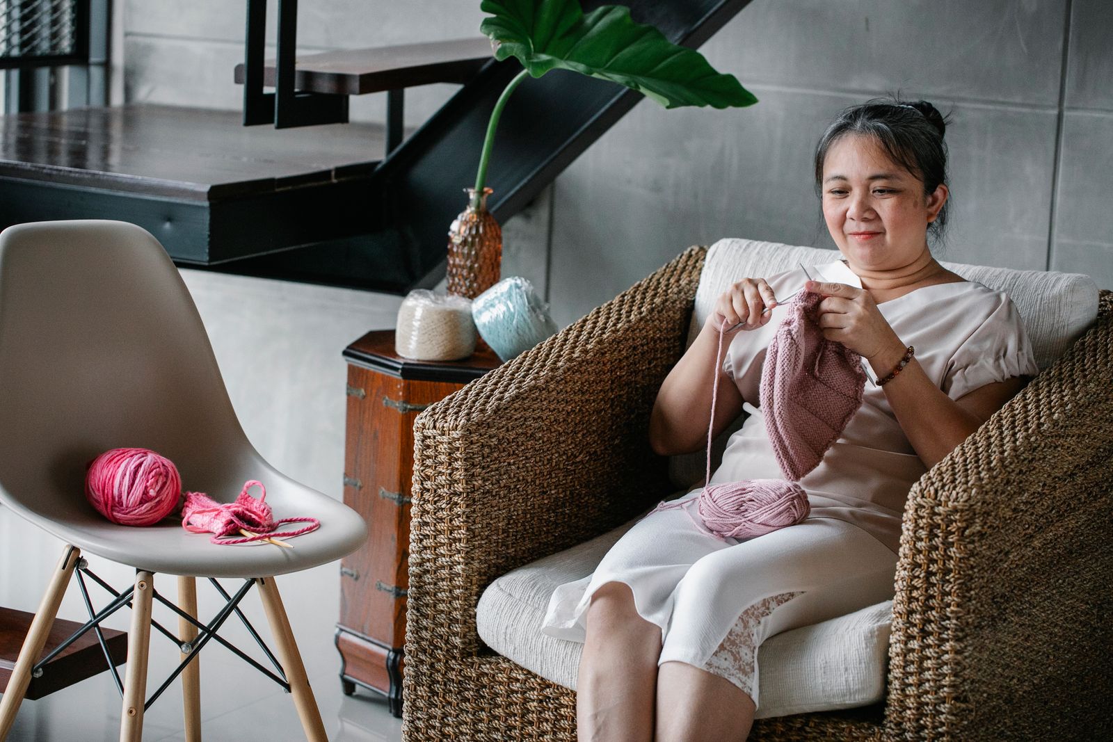 Content mature Asian lady in cozy rattan chair and knitting with needles and pink yarn