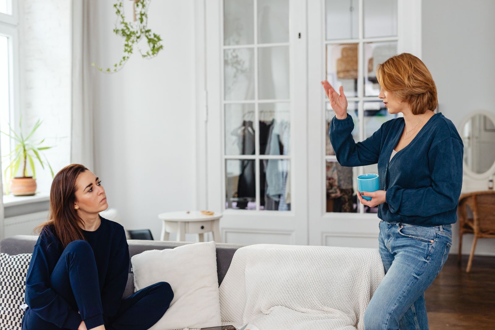 Two women engage in a friendly chat in a stylish living room, one sitting and one standing with a mug.