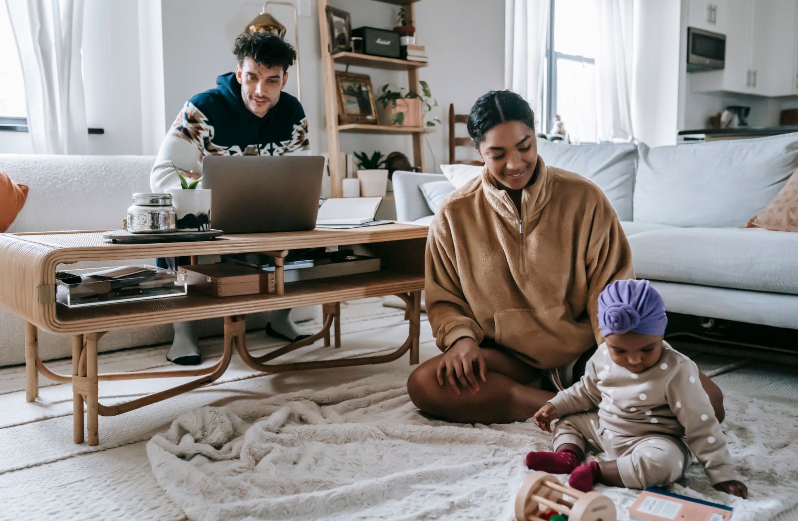 Focused ethnic father browsing laptop while working online at home with cheerful African American mother sitting on rug with little daughter