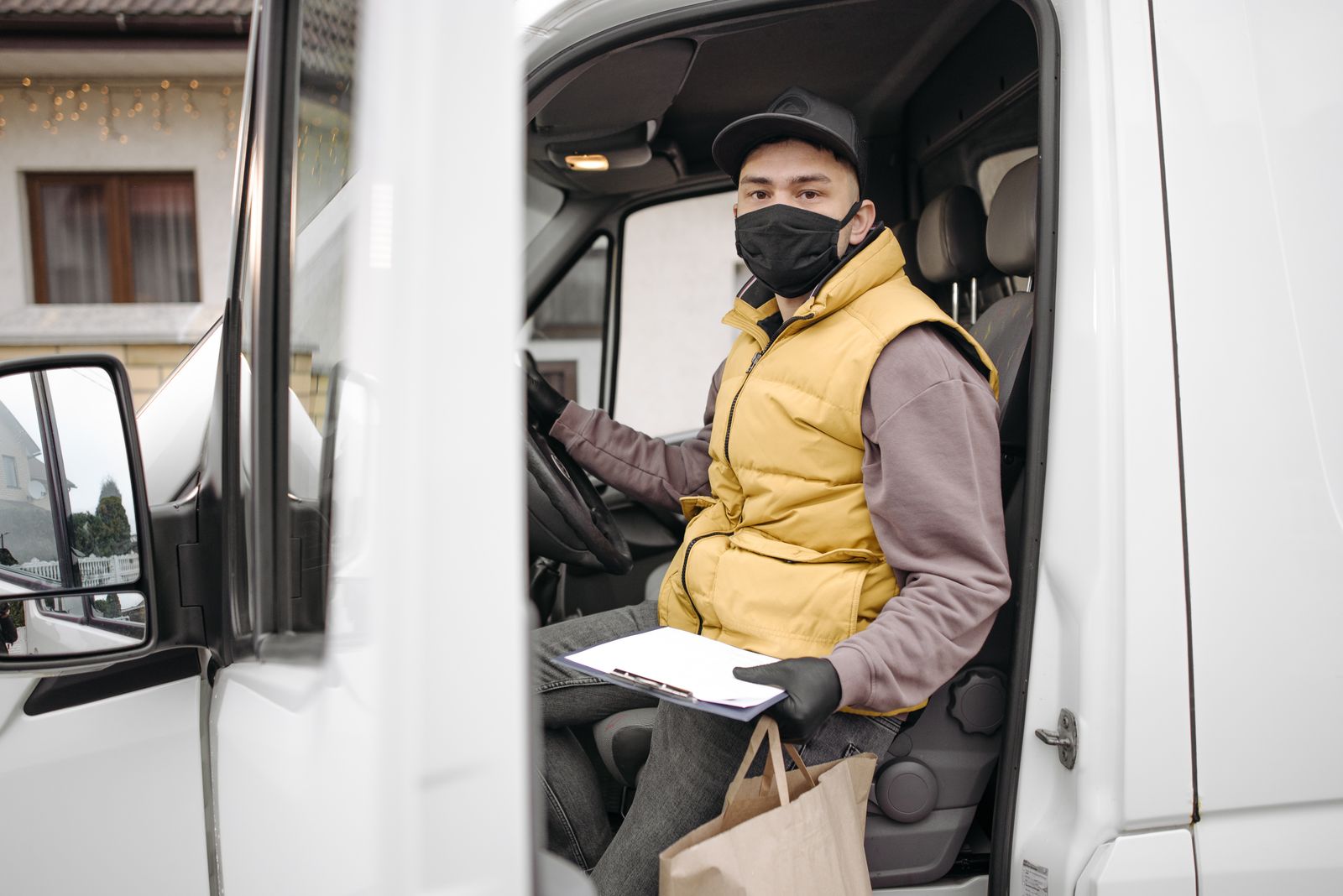 Delivery person in van wearing a facemask, holding clipboard and paper bag, during pandemic.