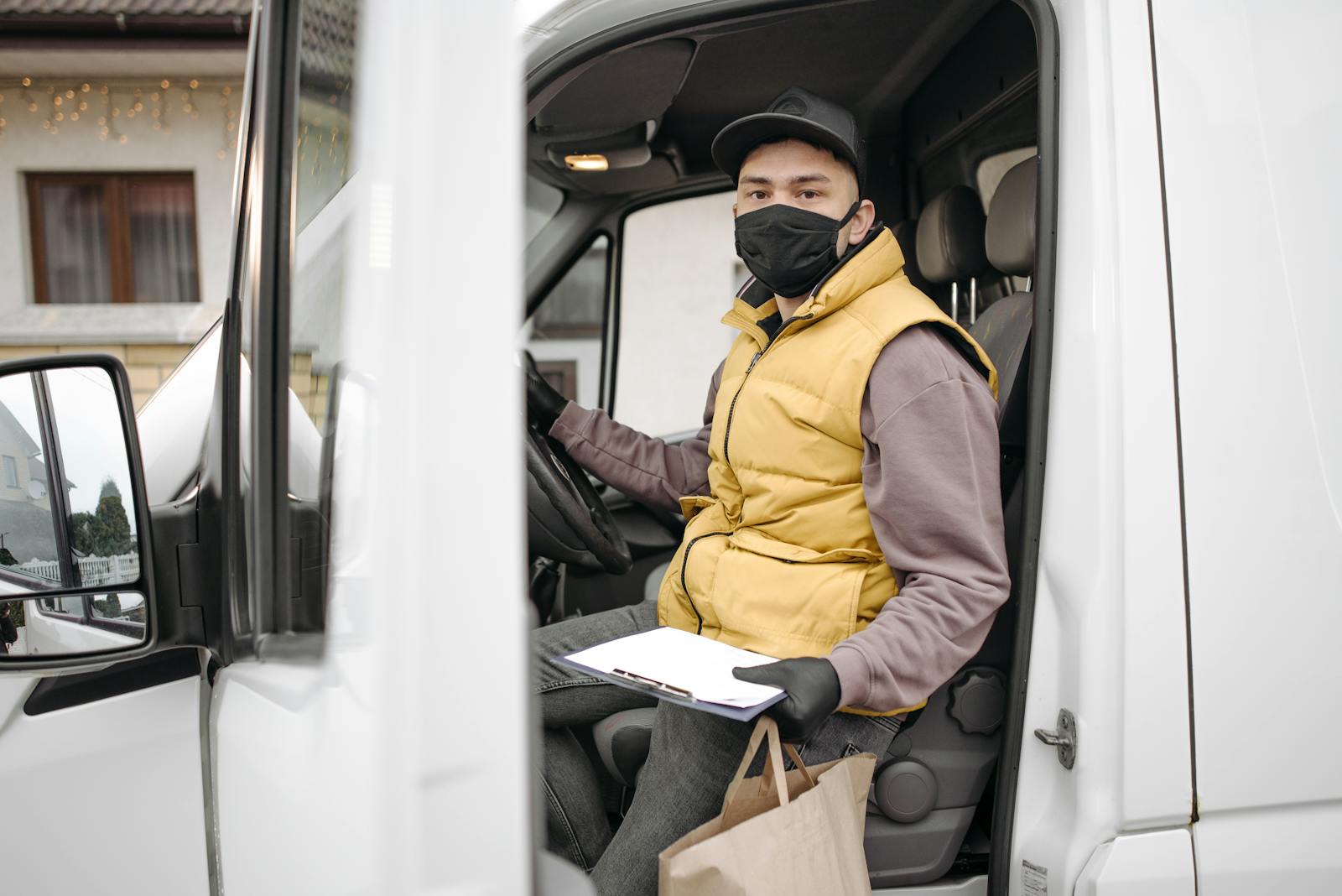 Delivery person in van wearing a facemask, holding clipboard and paper bag, during pandemic.