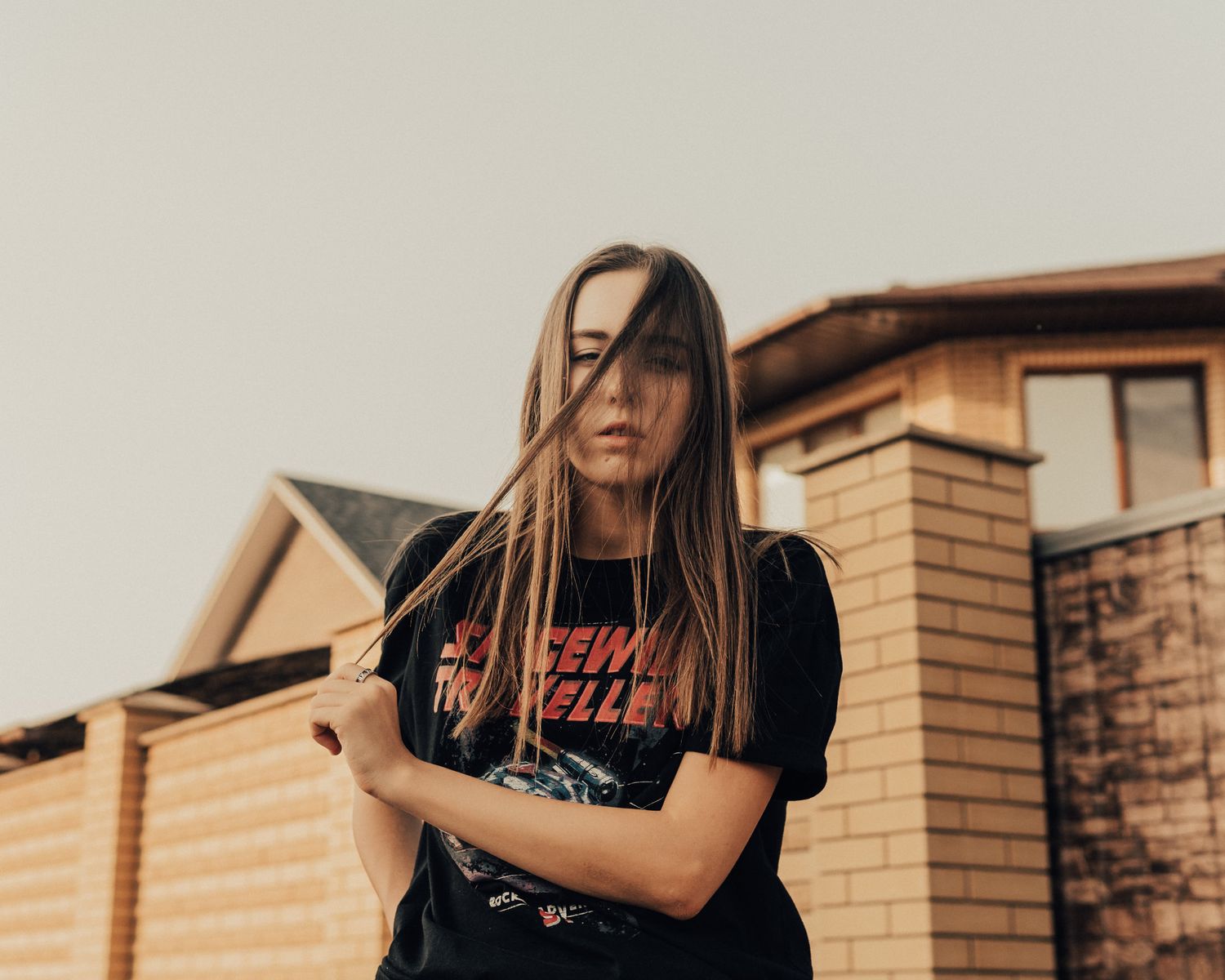 Serious female in black t shirt with hair covering face looking at camera while standing on street near house with stone fence