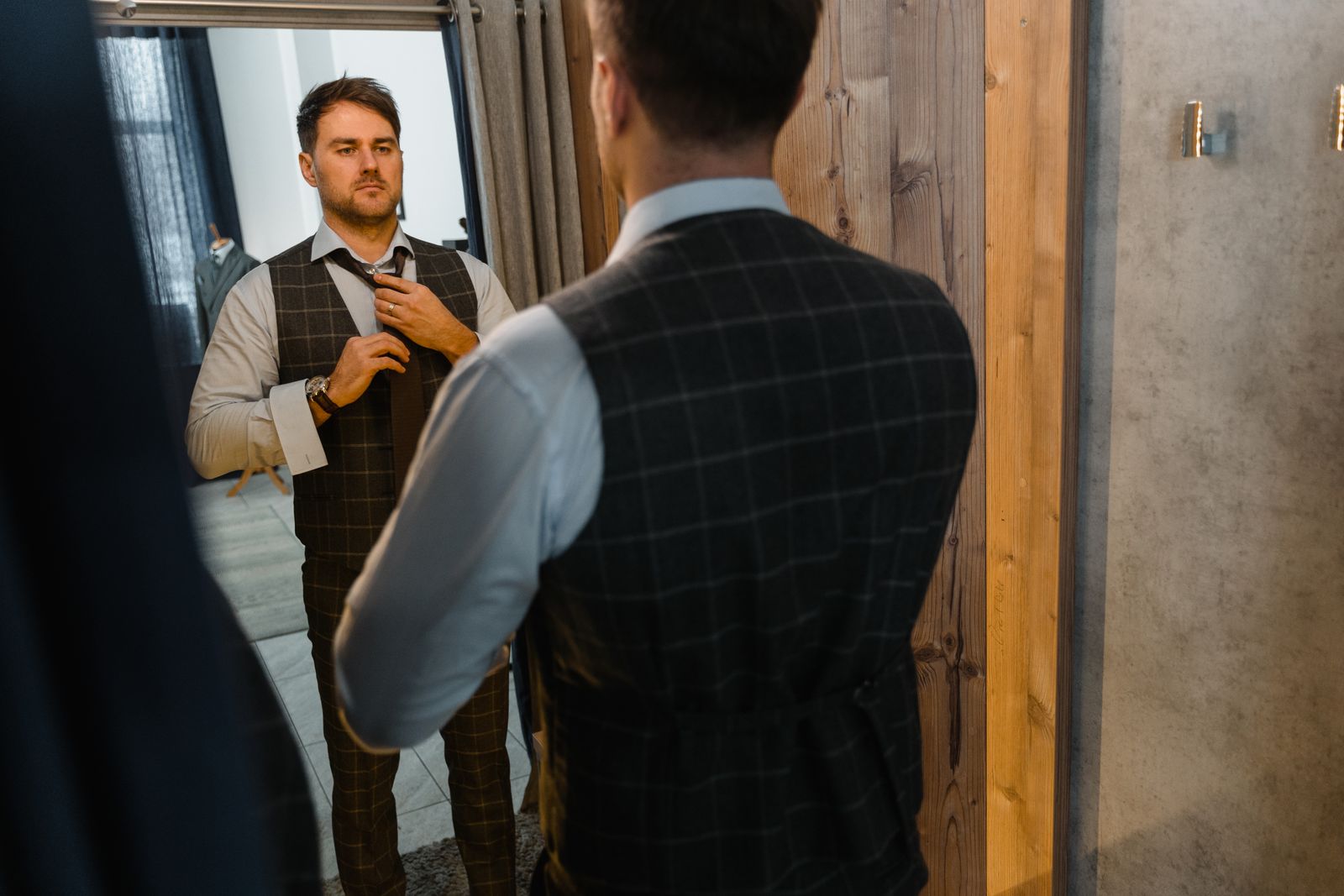Adult man in a stylish waistcoat adjusting his necktie while looking in the mirror indoors.
