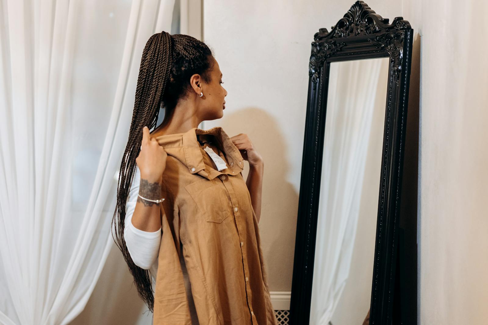 A woman with braided hair examines a dress in front of an ornate mirror indoors.