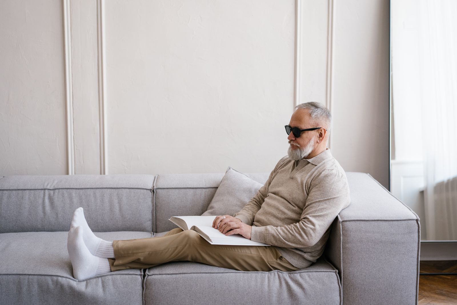 An elderly blind man reading a Braille book while sitting comfortably on a sofa indoors.