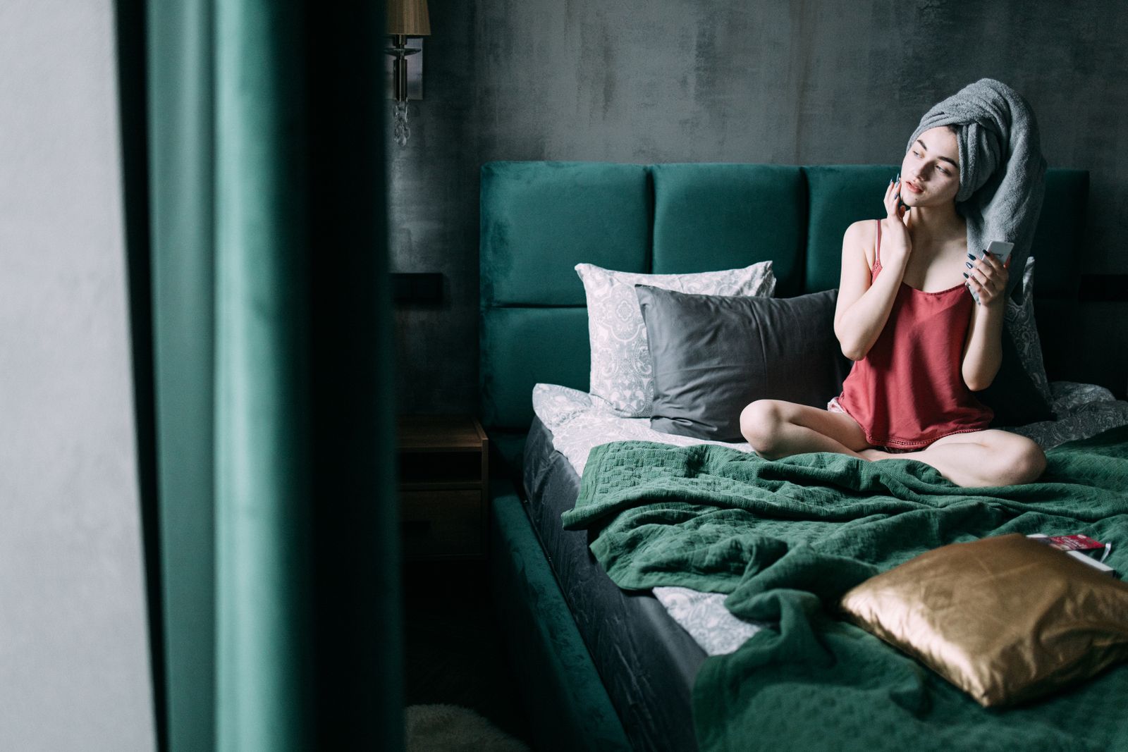 Caucasian woman moisturizes her face sitting on a bed, wrapped in a towel.
