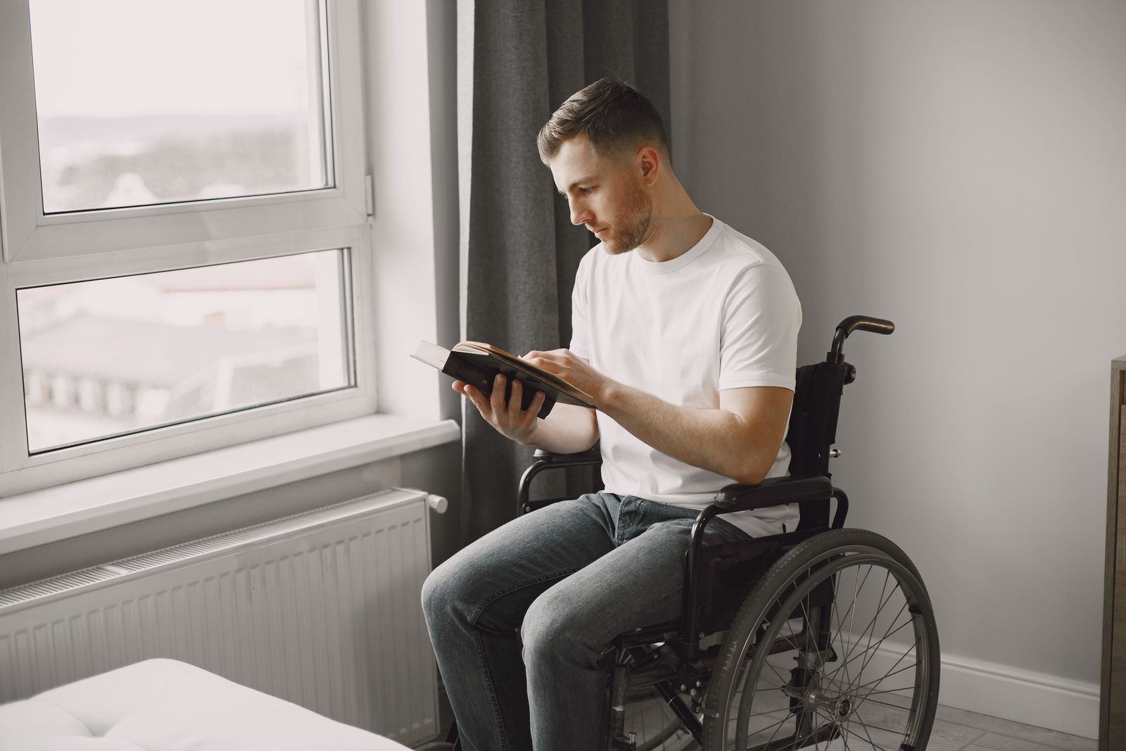 A man in a wheelchair reading a book in a well-lit room near a window.