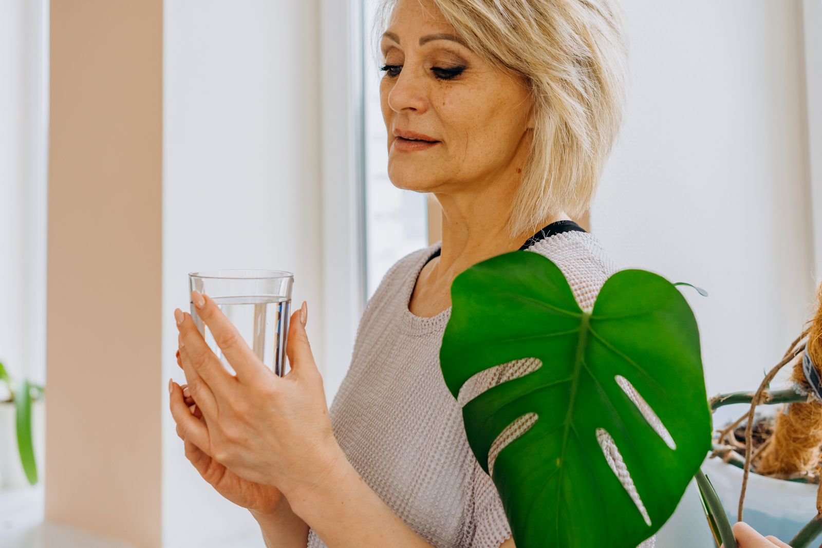 A serene senior woman holding a glass of water, standing near a bright window.