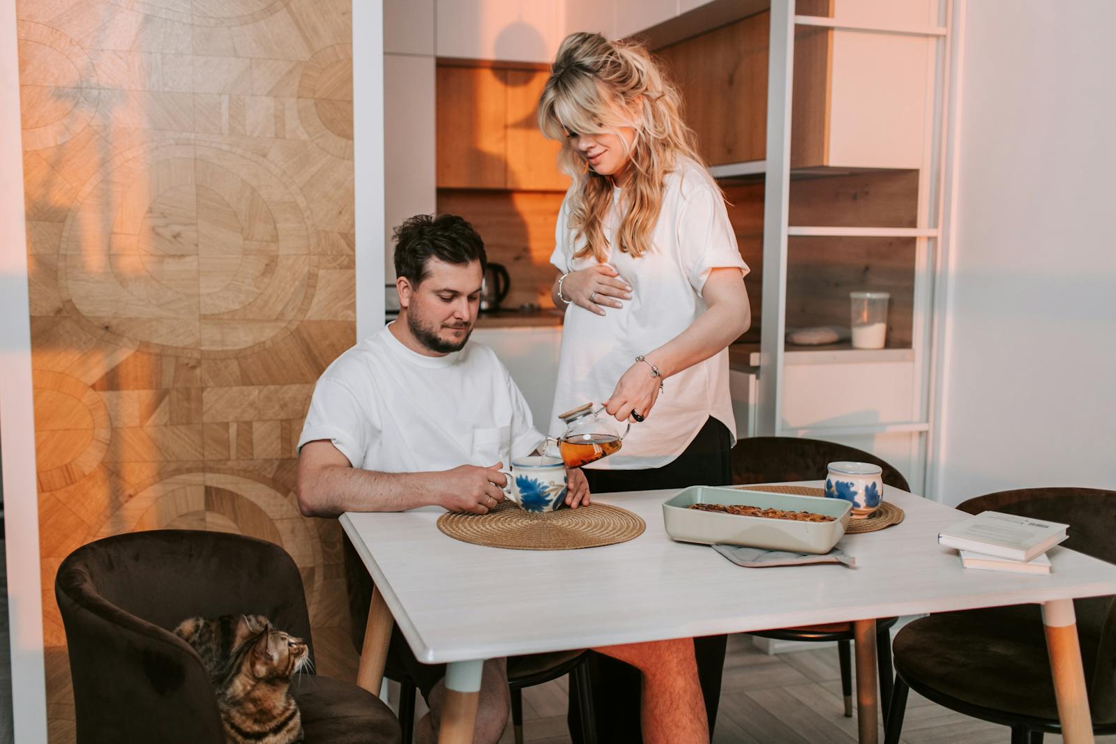 A pregnant woman and her partner sharing a peaceful afternoon with tea and a cat.