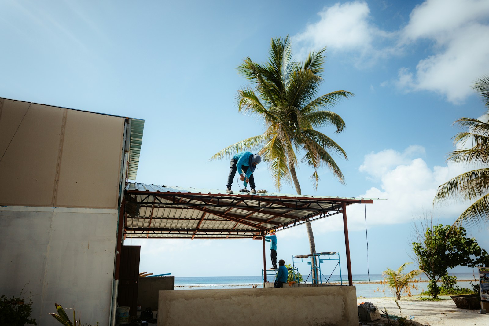 Workers repairing a roof near a beach with palm trees.
