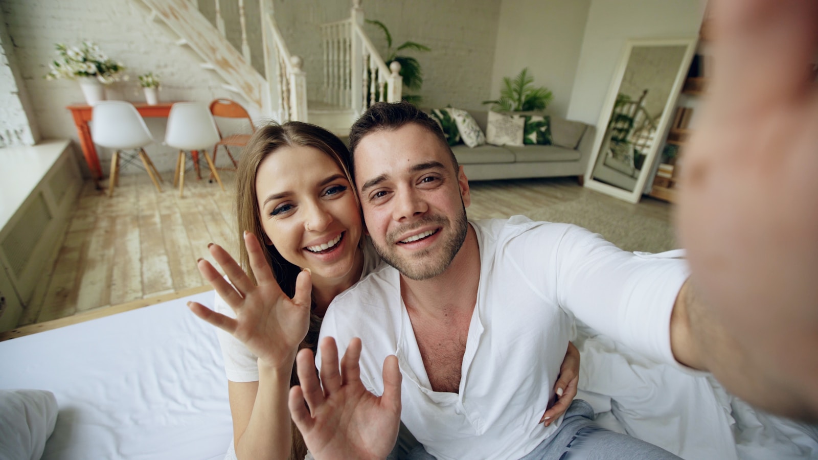 A happy couple waves hello from their bed.
