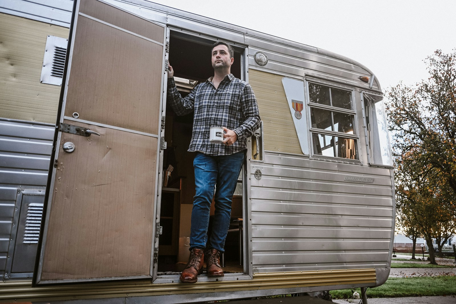 A man exits a vintage camper holding a coffee mug.