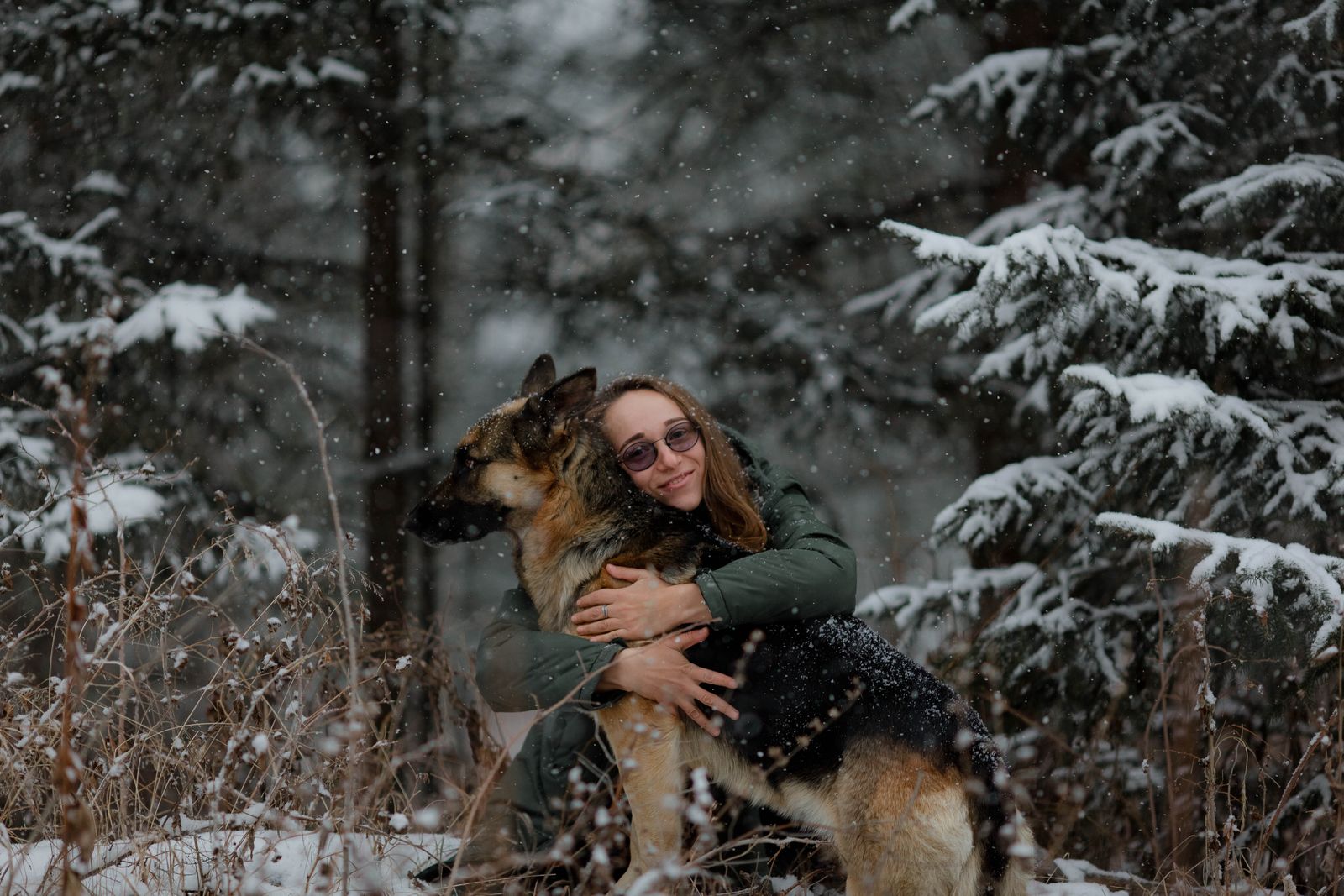 A woman embraces her German Shepherd dog amidst falling snow in a wintry forest setting.