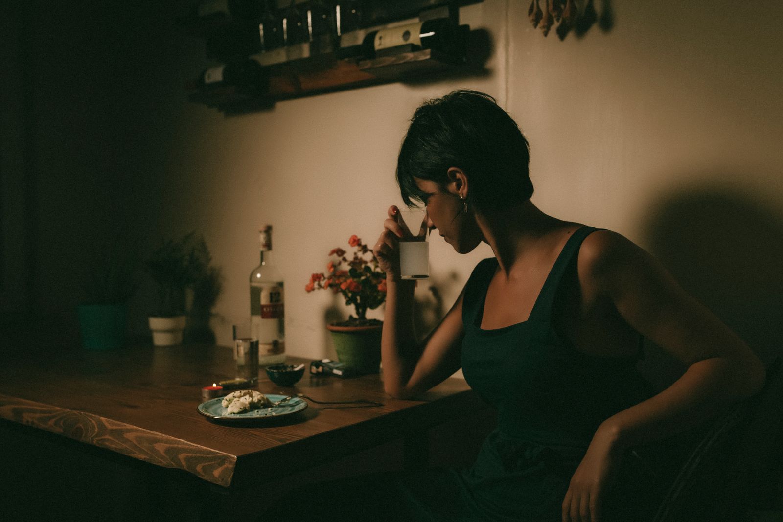 A solitary woman in a dimly lit room sits at a table, holding a cup as she contemplates her meal.