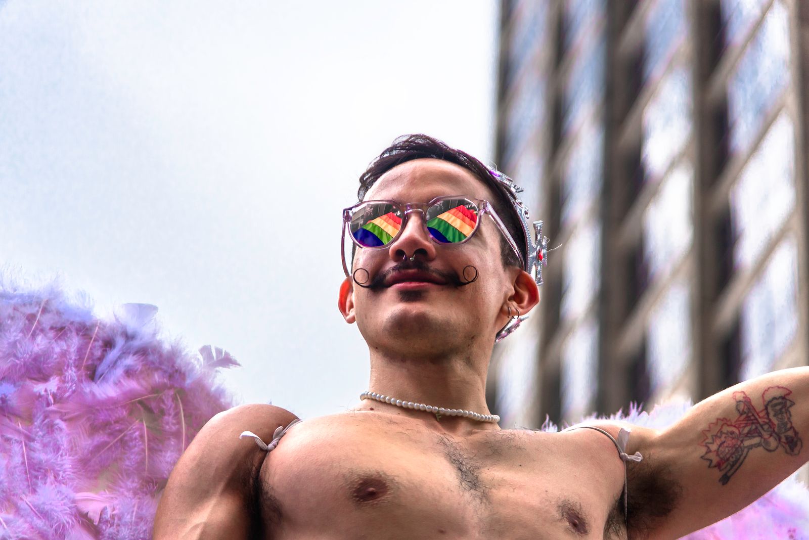 Man with rainbow sunglasses and feathers celebrates at an outdoor pride parade.