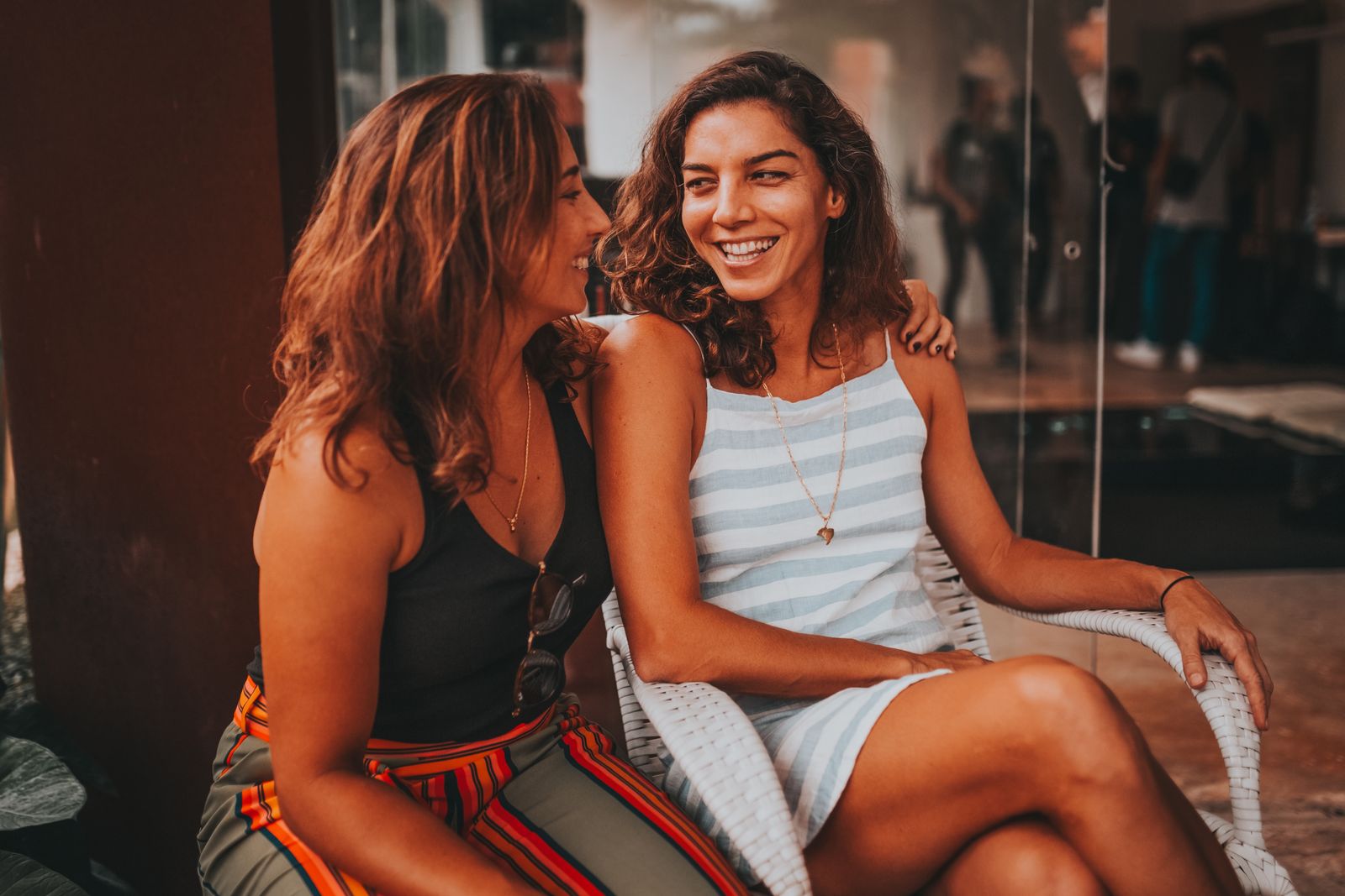 Two women sitting and smiling while chatting in a relaxed outdoor setting. Warm and friendly atmosphere.