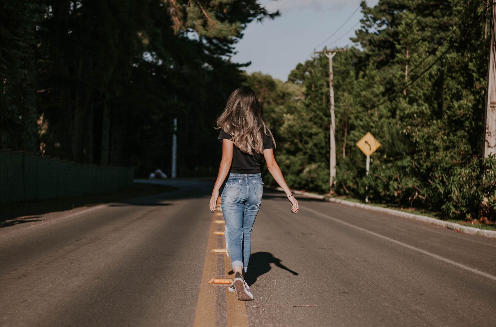 A woman casually walks down a sunlit road flanked by greenery, captured from behind during summer.