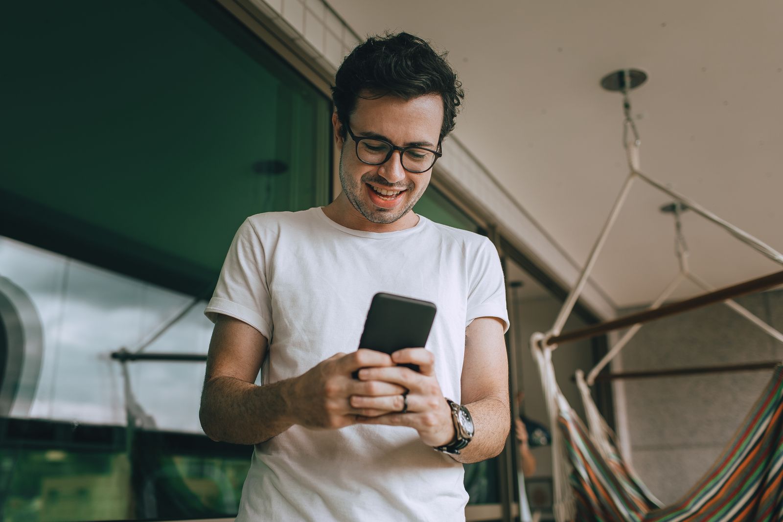 A cheerful young man with glasses checking his smartphone on a balcony.