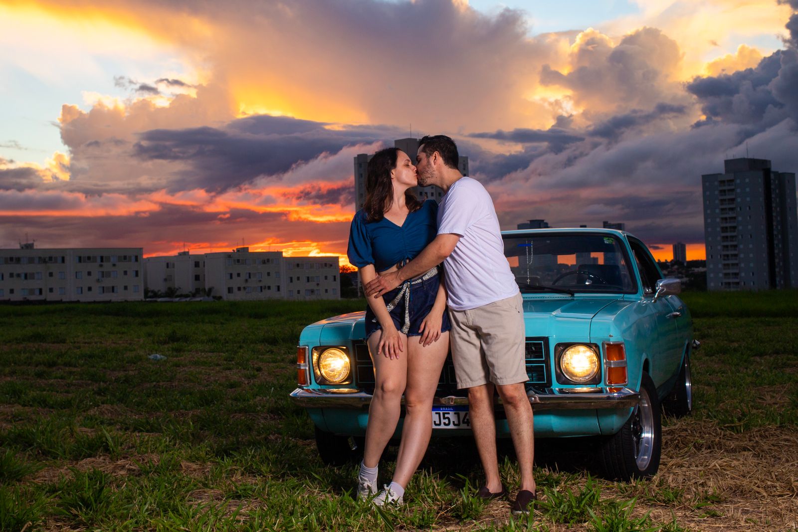 A couple embraces near a vintage blue car at sunset with a vibrant sky backdrop.