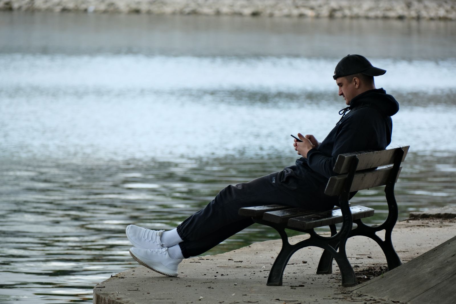 Casual young man seated on a park bench by a tranquil lake using a smartphone.