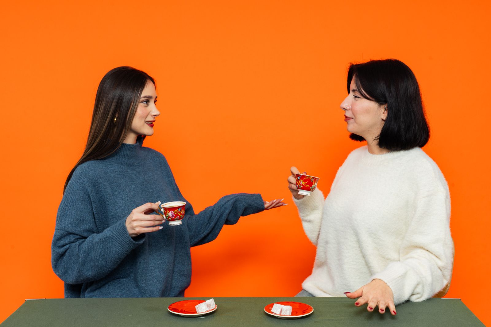 Two women having a conversation while drinking tea against an orange backdrop.
