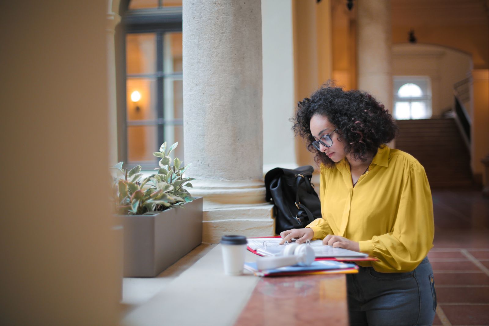 A young woman with curly hair studying in a bright corridor, wearing glasses and focusing on her work.