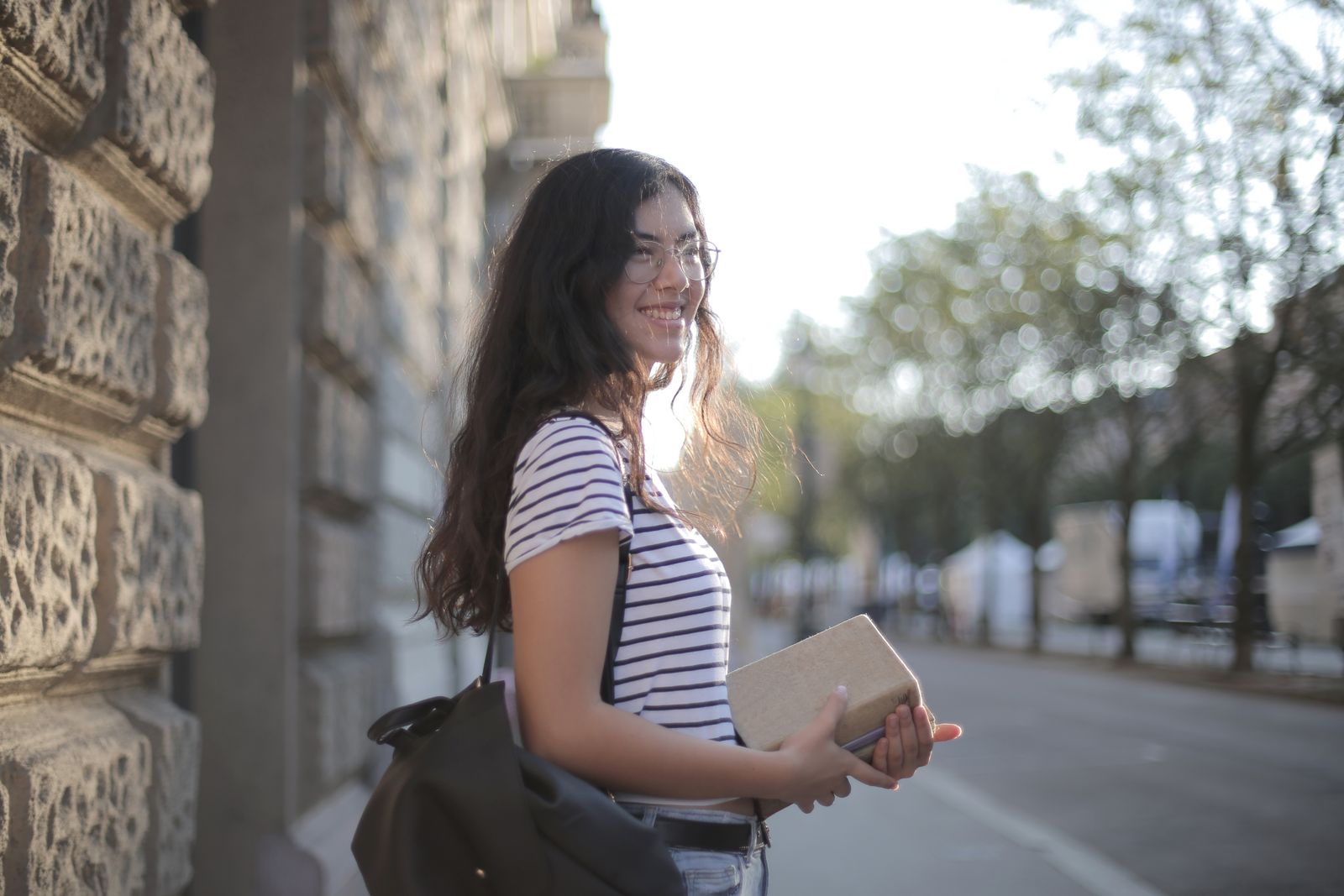 Side view of satisfied ethnic female wearing casual t shirt standing with pile of books and backpack on street while enjoying weekend and looking away