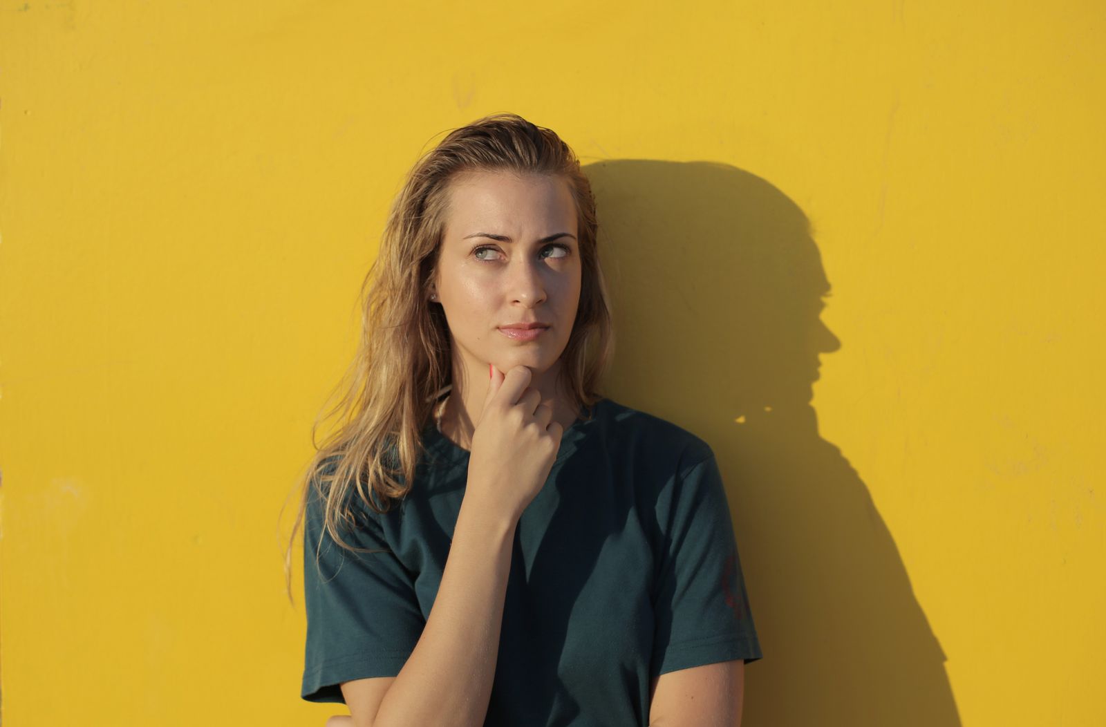 A thoughtful woman poses against a vibrant yellow wall, casting a shadow.