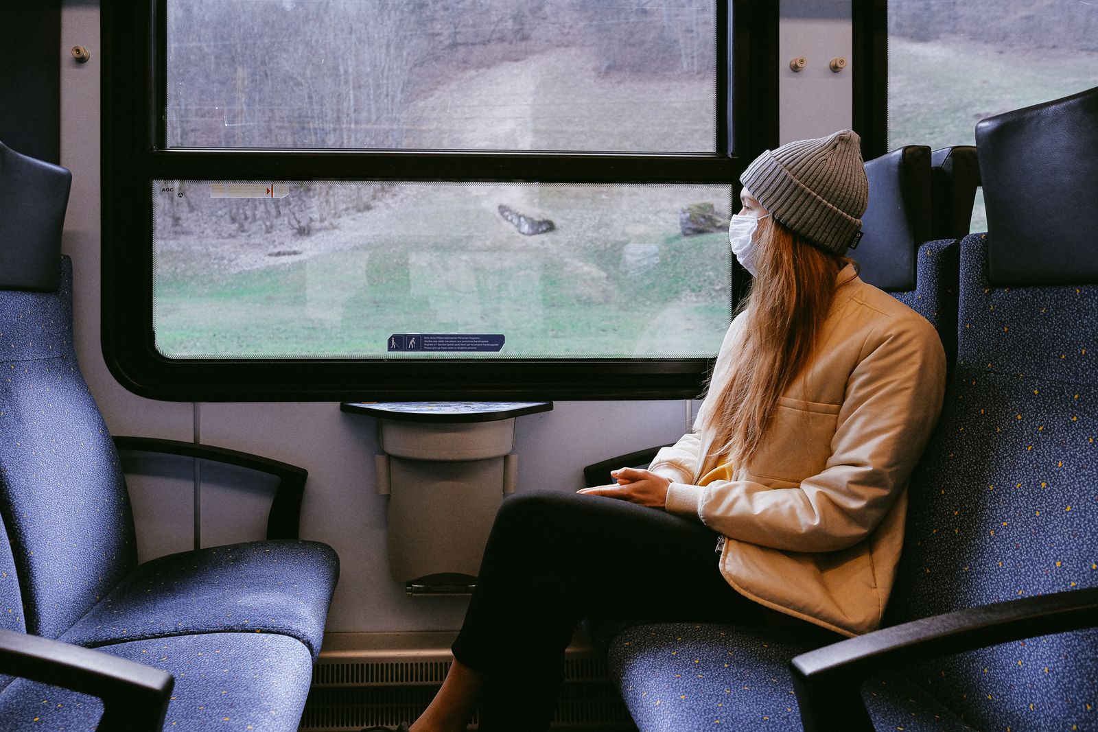 Woman wearing mask on train during pandemic, observing outdoor scenery.