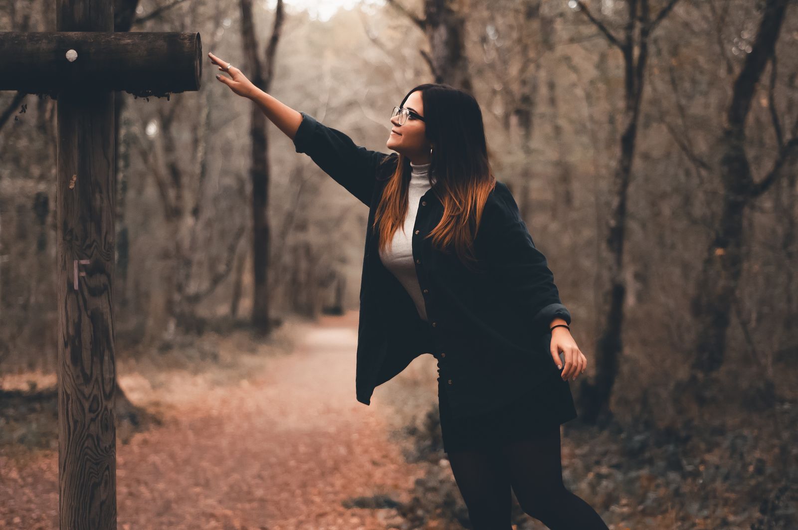 A stylish woman reaching out playfully while walking through a scenic autumn forest path.