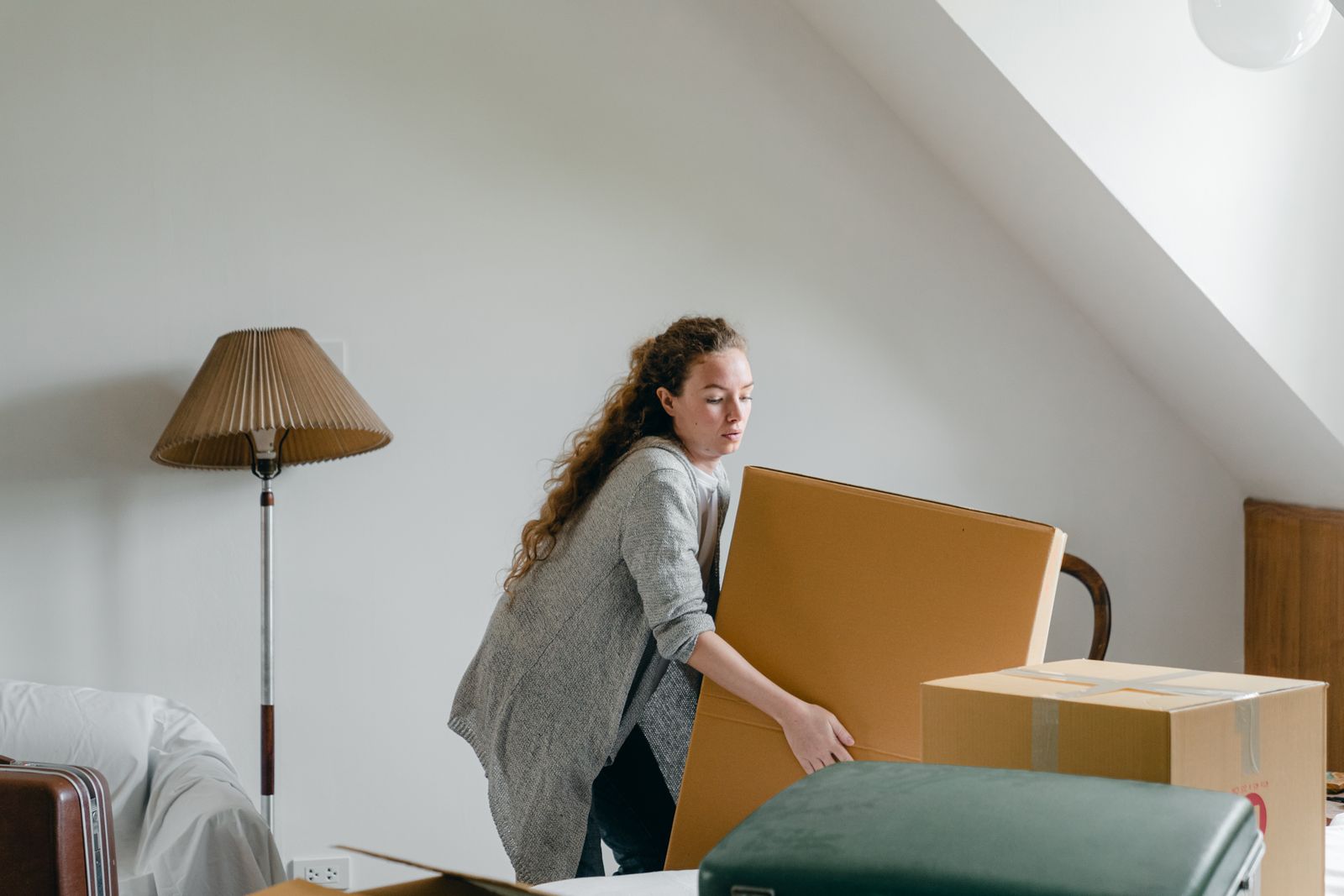 Slim female in cardigan taking big rectangular cardboard box while standing near suitcases and sofa in attic style living room in daylight