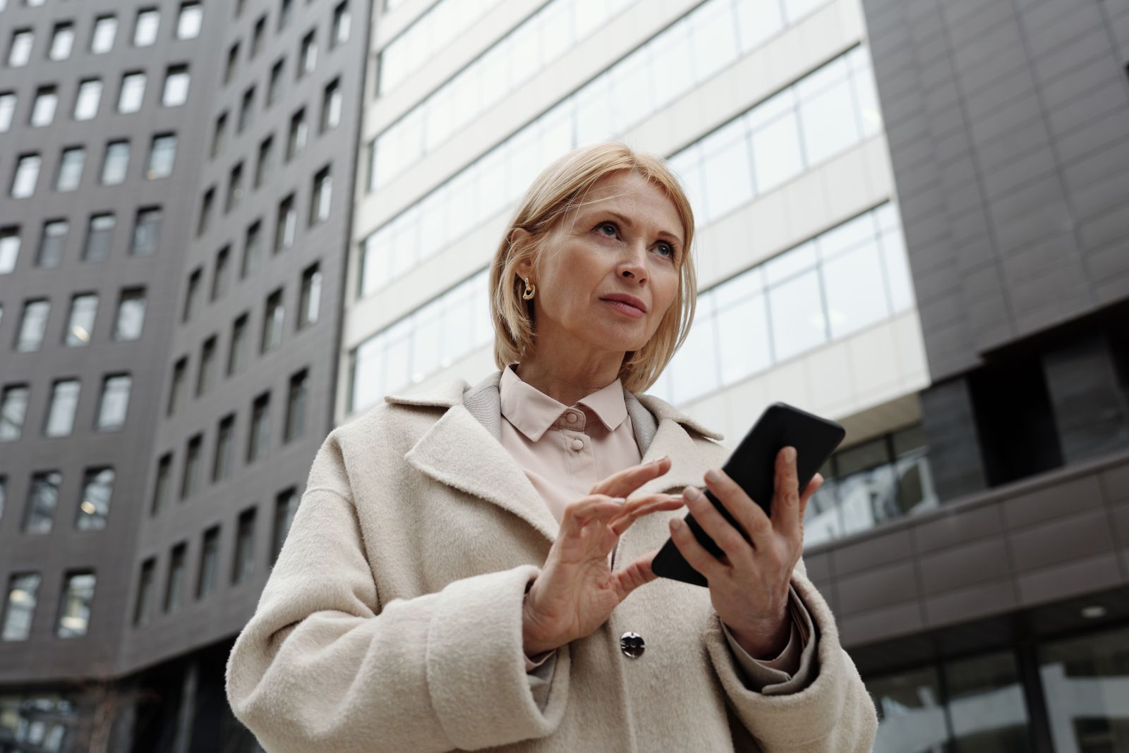 Businesswoman in a coat using a smartphone outside office buildings.
